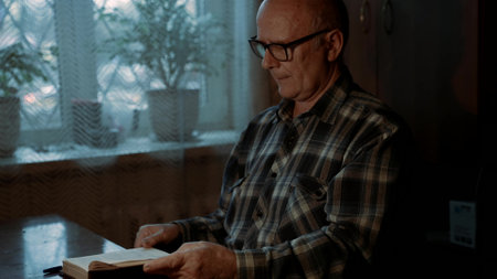 grandfather with glasses is reading book at home, sitting at table