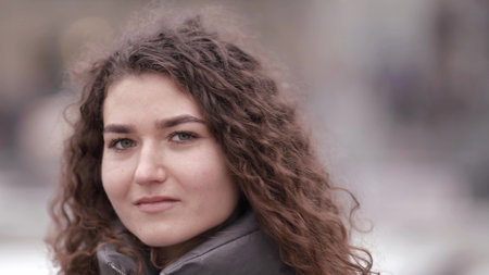 Close-up portrait of a young woman with curly hair posing on a city street, wearing a jacket, cold season.の写真素材