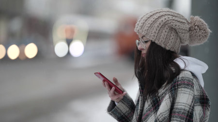 A beautiful young dark-haired woman in round glasses and a plaid coat walks around the city on a snowy winter day and looks at her smartphone, smiles, laughs, people pass by.の写真素材