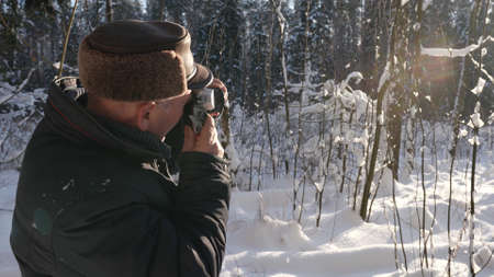 An elderly man takes photos of the winter sunny nature with a camera in the forest. There are trees...