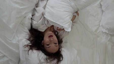 A brunette woman in a white shirt poses in a room on a bed on white bed linen. Top view.の写真素材