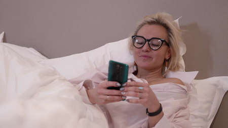 Close-up of a blonde woman with glasses is in the bedroom, lying on the bed with white rumpled bed linen and looking at her smartphone and laughing.の写真素材