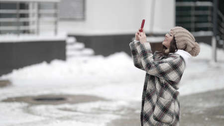 A young dark-haired woman in round glasses and a knitted hat is outside on a winter day, taking pictures of something on her smartphone and smiling.の写真素材
