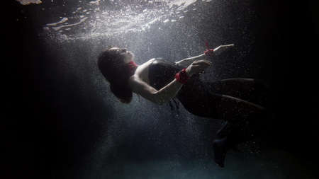 A dark-haired woman in red handcuffs swims deep under the water on a dark background, she soars and creates a lot of bubbles.の写真素材