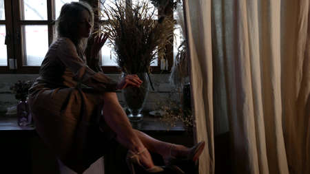 romantic adult woman is resting at home, sitting on windowsill and stroking her leg