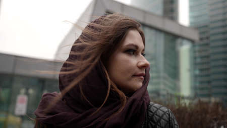 pretty young woman with scarf on head is standing on street of city at cold day, closeup female...