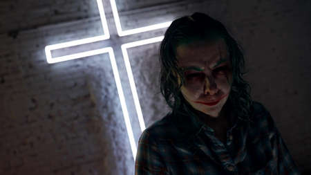 horror movie, person with clown makeup is standing against illuminating catholic cross