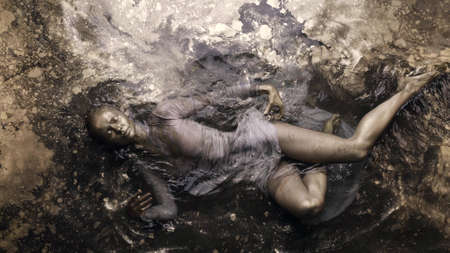 A woman in a dress with dyed golden skin and hair lies in the water, slowly moving and posing.