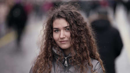 A close-up portrait of a young woman with curly hair standing on a city street on a cold day against the background of people passing by.の写真素材