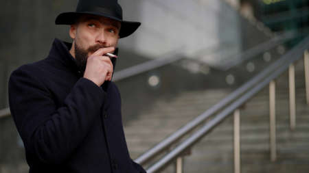suspicious man in black coat and hat is smoking cigarette on street and waiting someoneの写真素材
