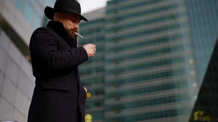 mysterious man in black coat and hat is lighting cigarette and smoking on street in modern cityの写真素材