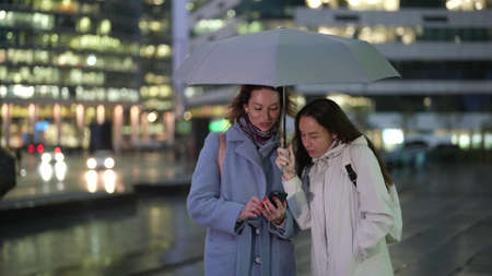 two adult women are meeting by chance on street in city in evening in rainy weather and chattingの写真素材