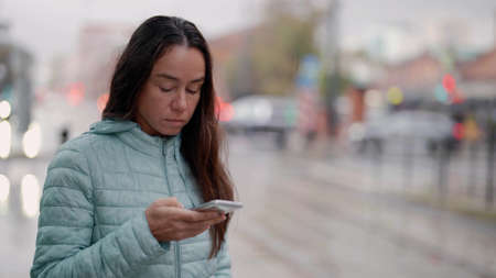 woman is waiting bus on station in city and sending message by smartphoneの写真素材