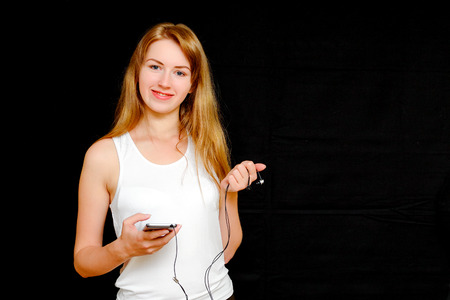 A smiling girl in a white shirt on a black background holding a smart phone and headphones.の写真素材