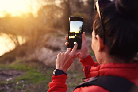 Girl taking landscape with river at sunset time photo with smartphone. Female tourist exploring new places.の写真素材