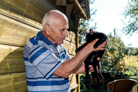 Elderly man holds pet black kitty in hands at home yard against yellow wooden wall outdoorの写真素材