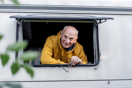 Positive elder man with glasses in hand looking through window of white motorhome in campingの写真素材