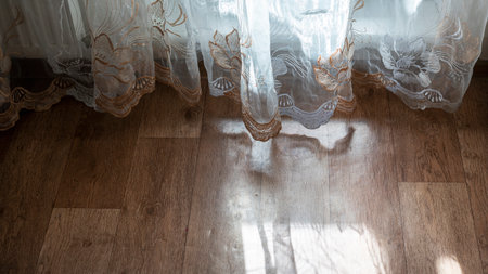 Sheer lace curtain with floral embroidery touch wooden floor while sunlight casts delicate shadows through fabric. Warm light highlights intricate patterns and adds cozy atmosphere to interior.の写真素材