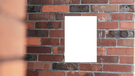 Blank white poster taped on red brick wall partially obstructed by another brick structure on left. Tape is slightly visible securing poster against weathered and uneven brick surface.の写真素材