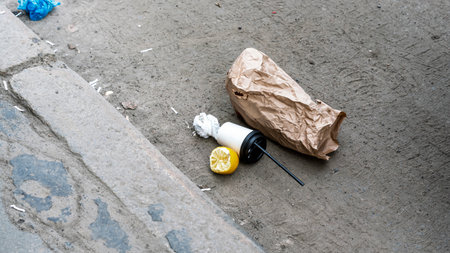 Litter scattered on concrete surface including crumpled brown paper bag, disposable cup with black...