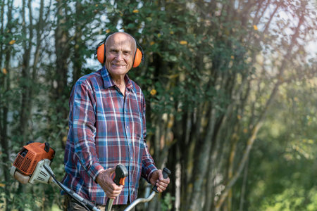 Elderly man in plaid shirt wearing ear protectors holding a grass trimmer outdoors surrounded by green trees on a sunny day.の写真素材