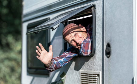 Mature man leaning out of caravan window smiling and waving his hand on bright sunny day.の写真素材