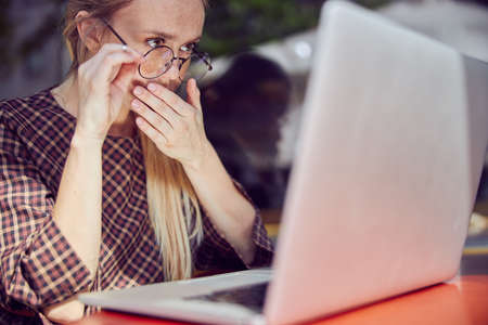 Waist up shot of beautiful red-haired girl with freckles in glasses who is sitting at the table and...