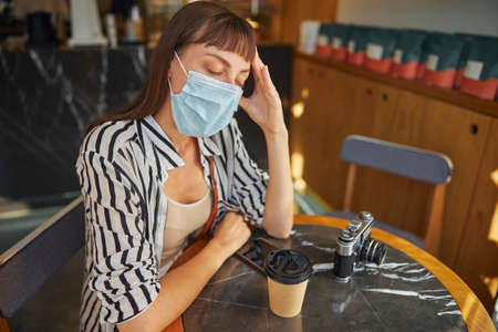 Young tired of work woman in mask is sitting with closing eyes in cafe