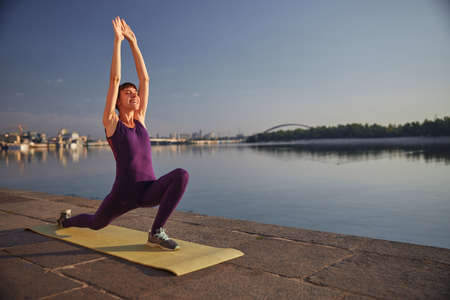 Young fun woman is doing Separate Leg Stretching Pose with hands up during morning yoga outdoor