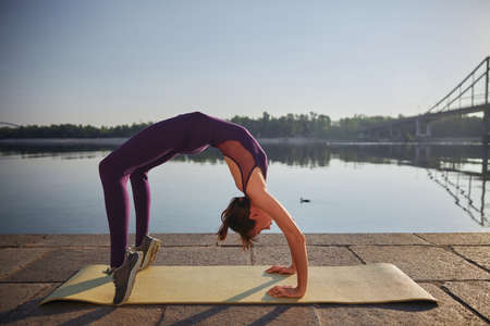A side shot of female who is doing yoga Wheel Pose near river
