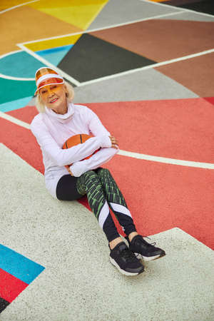 Old, healthy and cheerful lady with basketball ball on a colorfull square outdoorsの写真素材