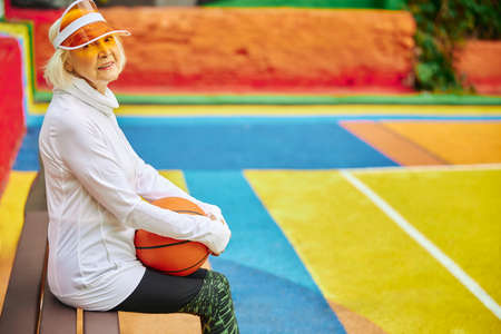 Old, healthy and cheerful lady with basketball ball on a colorfull square outdoorsの写真素材