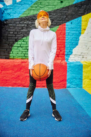 Old, healthy and cheerful lady with basketball ball on a colorfull square outdoorsの写真素材