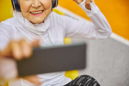 Old, healthy and cheerful lady on a colorfull square outdoors wearing headphonesand holding...