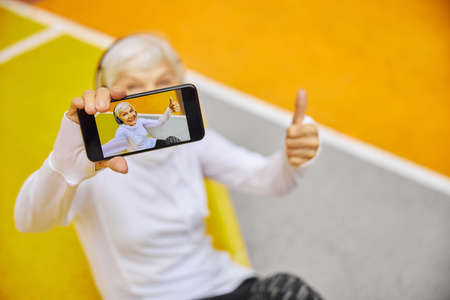 Old, healthy and cheerful lady on a colorfull square outdoors wearing headphonesand holding smartphoneの写真素材