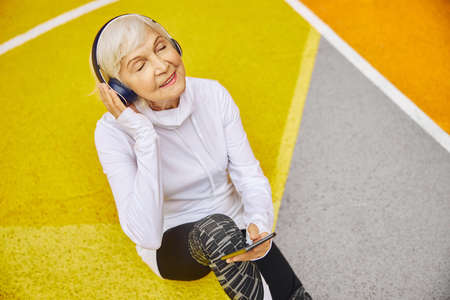 Old, healthy and cheerful lady on a colorfull square outdoors wearing headphonesand and listening to musicの写真素材