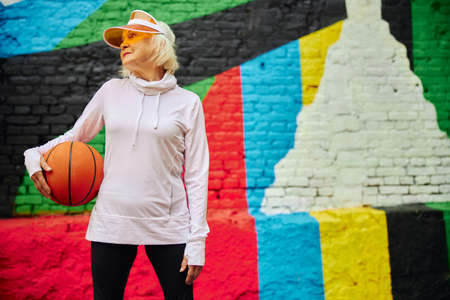 Old, healthy and cheerful lady with basketball ball on a colorfull square outdoorsの写真素材