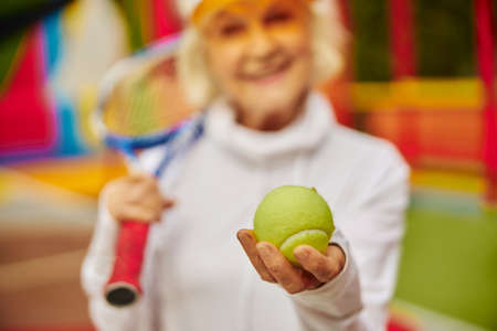Old, healthy and cheerful lady on a colorfull square outdoors and holding tennis racket with ballの写真素材