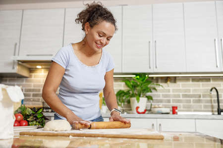 Beautiful smily handsome woman is preparing tasty fresh italian pizzawith vegetables at her kitchen...