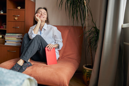 Young cheerful confident and happy woman is relaxing at her cozy armchair at home with booksの写真素材