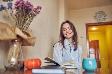 Young cheerful confident and happy woman is enjoying tea while reading book at her kitchen at homeの写真素材