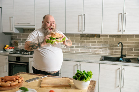 Emotional fat funny and tattooed man with food at the kitchen at homeの写真素材