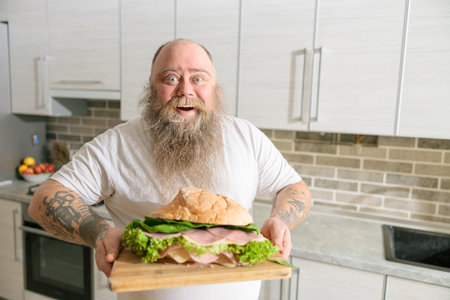 Emotional fat funny and tattooed man with food at the kitchen at homeの写真素材