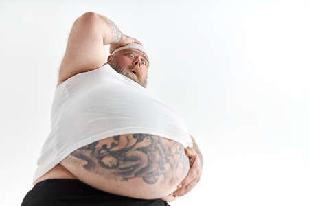 fat man with big belly and tattoes in sports wear is holding his stomach with shocked emotionの写真素材