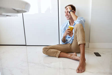 Caucasian hadsome young man in blue shirt is sitting on the floor and chilling at modern kitchen