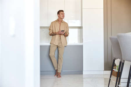 Hadsome smiling man is chilling on the minimalistic kitchen with smartphoneの写真素材