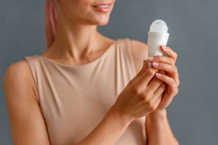 Close up shot of beautiful young woman holding antiperspirant and smiling isolated over gray studio...