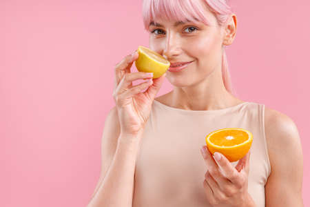 Portrait of smiling woman with pink hair holding half of ripe orange and smelling fresh lemon, posing isolated over pink backgroundの写真素材