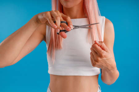 Cropped shot of woman with pink hair cutting her hair using scissors, standing isolated over blue studio backgroundの写真素材