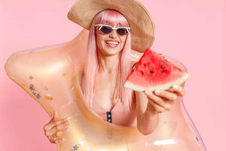 Excited woman with pink hair in swimsuit and sunglasses holding watermelon slice and inflatable star for swimming isolated over pink backgroundの写真素材